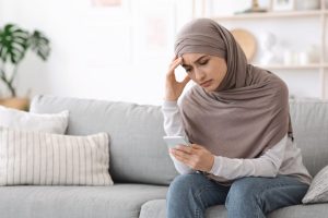 A young woman in a hijab sitting on a sofa holding her head, a phone in her hand