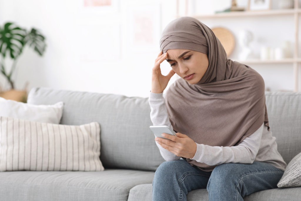 A young woman in a hijab sitting on a sofa holding her head, a phone in her hand