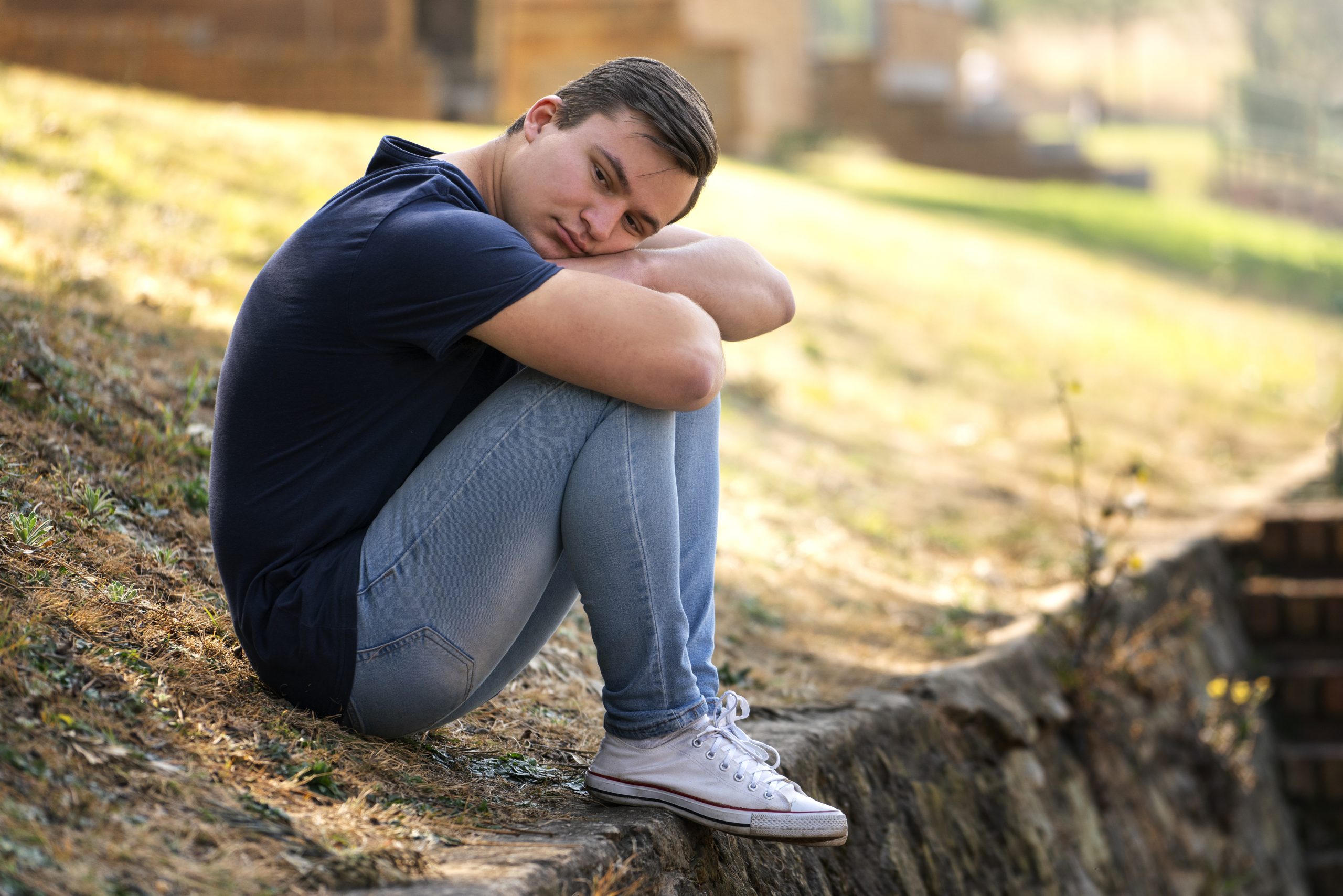 A young man sitting outdoors with his arms around his knees, looking down