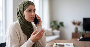 A young Muslim woman in a hijab sits at her kitchen table talking on her mobile phone, looking worried.