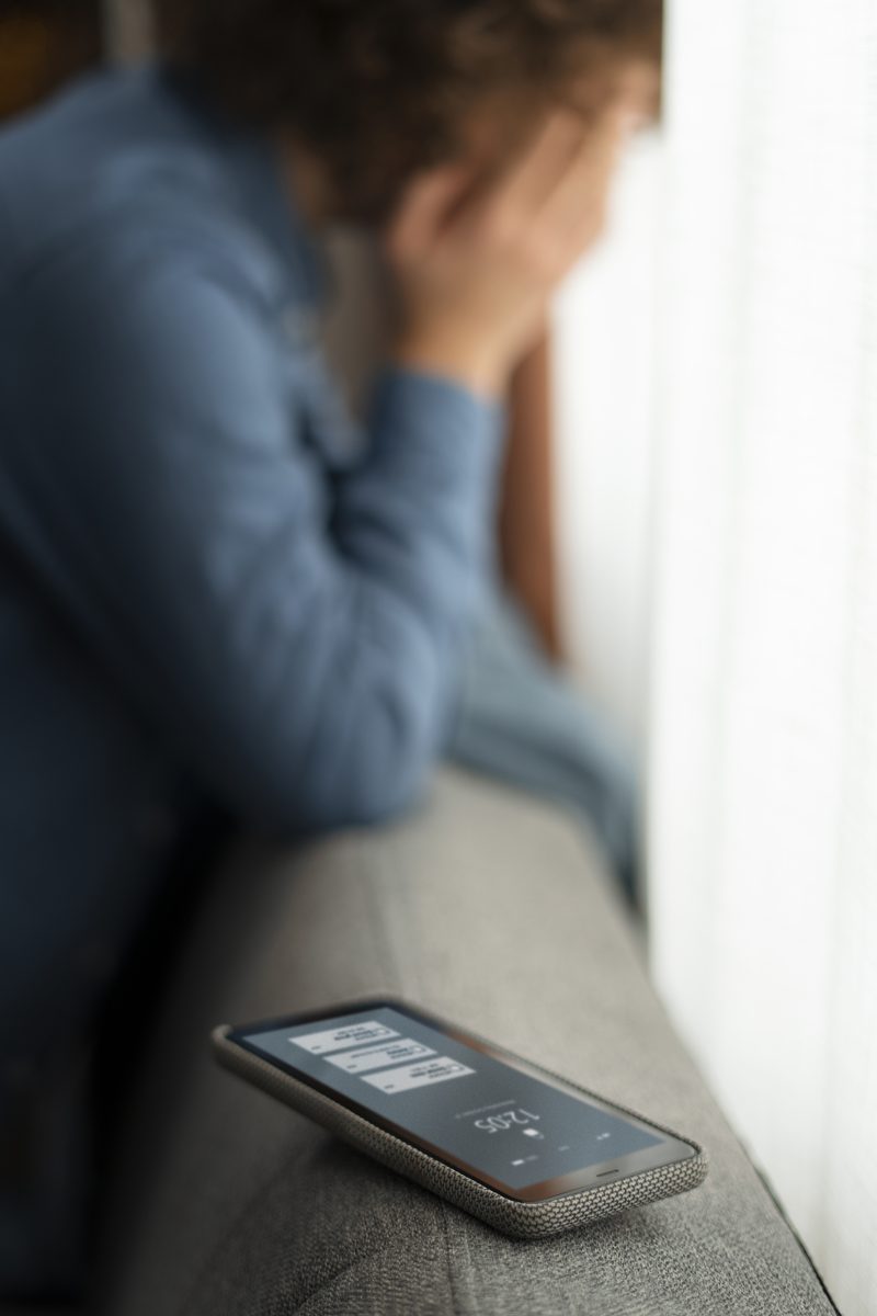 A young person sits alone by a window with their head in their hands, a phone face up on the sofa beside them.
